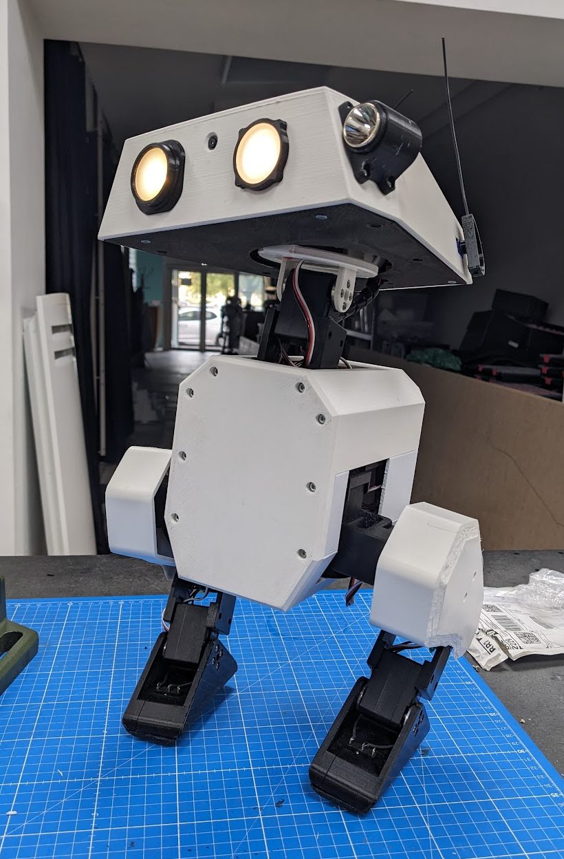 Front view of an assembled Open Duck Mini v2 bipedal robot with white 3D-printed body, glowing eyes and black servo legs, standing on a blue cutting mat